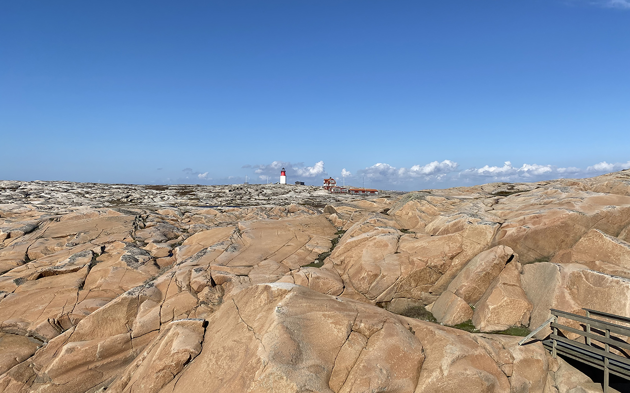 Vidsträckta ljusa klipphällar med en vit fyr med röd topp och röda byggnader under klarblå himmel